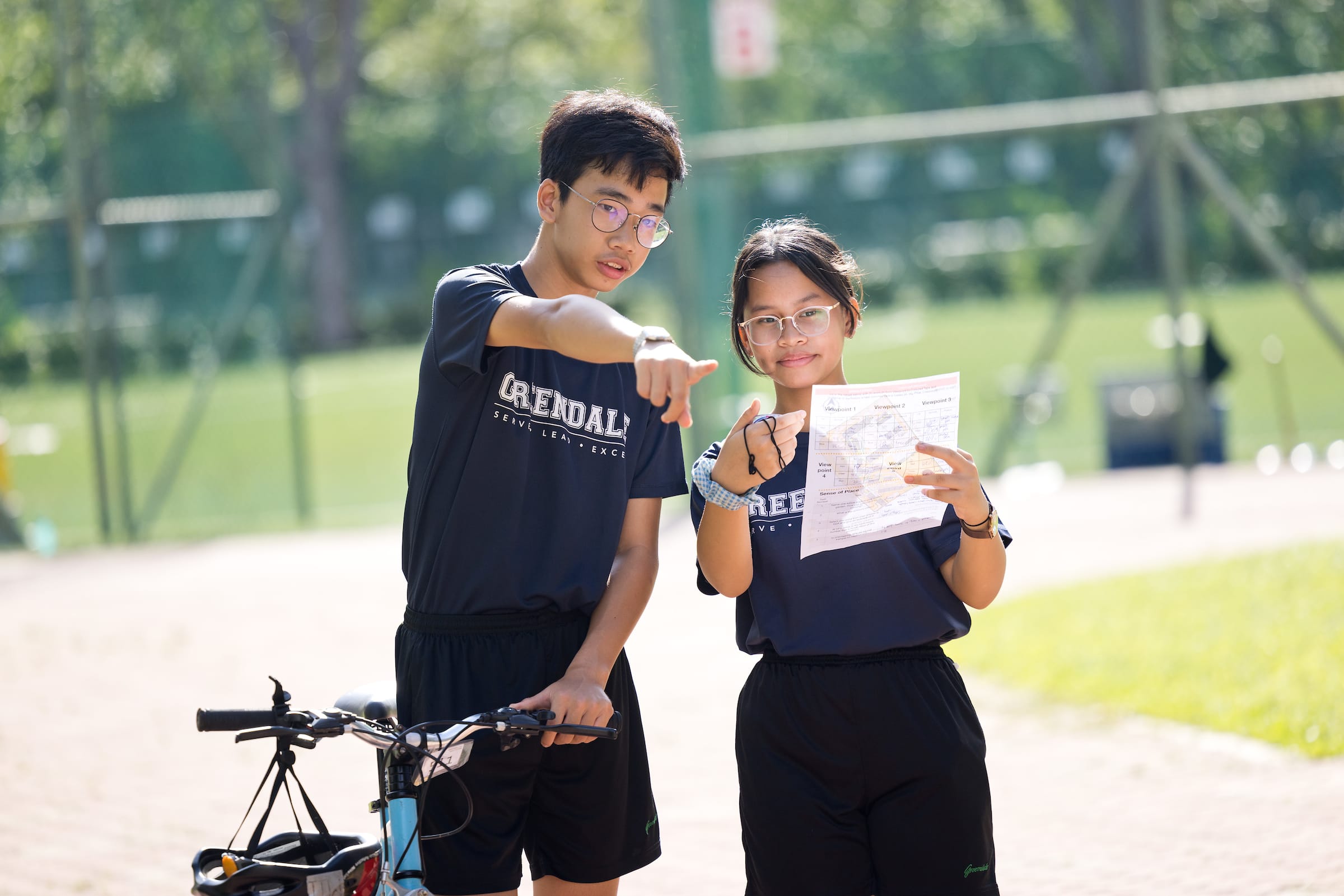 wo students standing outdoors with a bicycle, one holding a map and the other pointing toward a distance to navigate.